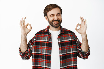 Smiling male model with beard, showing ok, okay signs and nod in approval, standing in checked...