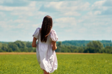 Fototapeta na wymiar A young woman in a white dress, against the background of a green field, stands with her back to the camera.