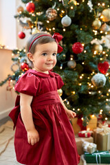 winter holidays and childhood concept - lovely baby girl in red dress over christmas tree at home