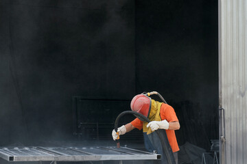 Worker in protective equipment using sandblaster for cleaning surface of metal sheet before...