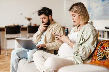 Man and pregnant woman using cellphone and laptop while sitting on couch