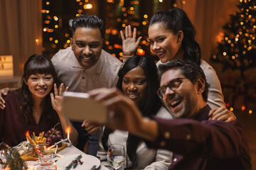 holidays, party and celebration concept - multiethnic group of happy friends having christmas dinner at home and taking selfie with smartphone and waving hands