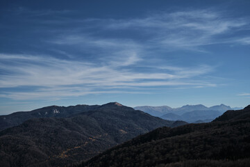 Beautiful panoramic landscape view of mountains on warm sunny autumn day. Blue sky and white clouds. In distance, steep cliffs and silhouettes of large snow capped mountain peaks.