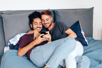 happy young gay couple lying on the bed watching a video on their mobile or making a video call or...