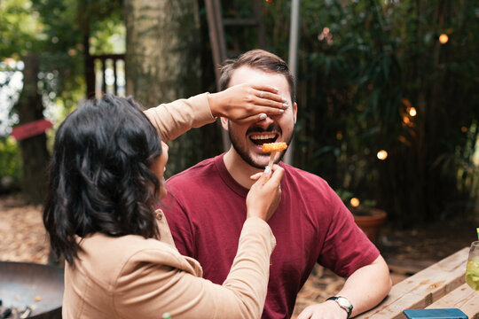 Two Friends Are Playing With Food Sitting At The Table In The Outdoor Restaurant - Young Couple In A Funny Moment - Blind Food Tasting