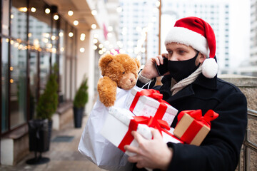 Man wearing face mask and Santa Claus hat holding christmas gift boxes after shopping in city mall....