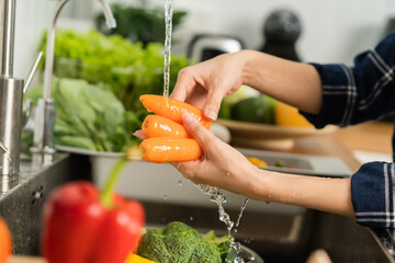 Asian young woman washing carrot, tomato, broccoli  fresh vegetables, paprika with splash water in basin of water on sink in kitchen, preparing fresh salad, cooking meal. Healthy food people.