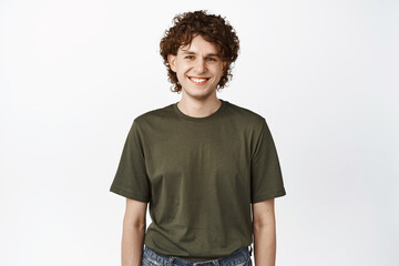 Portrait of handsome smiling curly young man, wearing green t-shirt, looking happy and relaxed at camera, standing over white background
