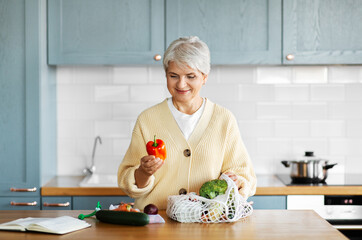 healthy eating, food cooking and culinary concept - happy smiling woman with vegetables in string bag and cook book on kitchen