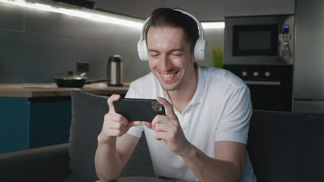 Smiling man watching funny mobile videos on his smartphone, while resting on sofa at home. Young male listening music with headphones and mobilphone, sitting on couch at home apartments