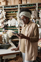 Confident African designer in eyeglasses reading information online on digital tablet while standing in the workshop