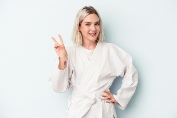 Fototapeta premium Young caucasian woman doing karate isolated on blue background joyful and carefree showing a peace symbol with fingers.