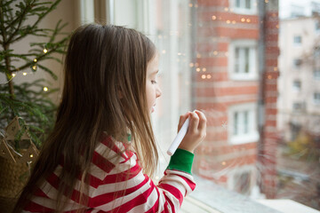 child draws snowflakes on window with chalk marker. girl in New Year's pajamas on windowsill against background of live Christmas tree in pot in wicker basket - obrazy, fototapety, plakaty