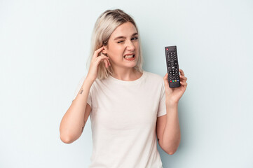 Young caucasian woman holding a tv controller isolated on blue background covering ears with hands.