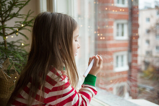 child draws snowflakes on window with chalk marker. girl in New Year's pajamas on windowsill against background of live Christmas tree in pot in wicker basket