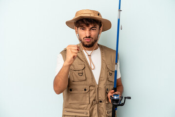 Young mixed race fisherman holding a rod isolated on blue background showing fist to camera, aggressive facial expression.