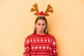 Young caucasian woman wearing a christmas reindeer hat isolated on yellow background shrugs shoulders and open eyes confused.