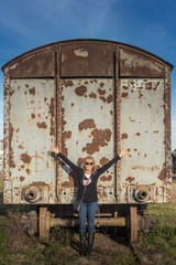 Adult woman wearing sunglasses smiling with her arms raised leaning against an abandoned train carriage.