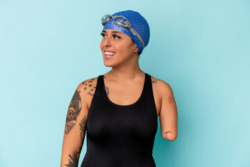 Young swimmer caucasian woman with one arm isolated on blue background looks aside smiling,...