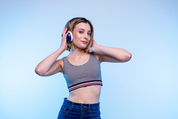 Young modern happy caucasian girl posing standing listening music with headphones with a light blue background - Studio photography of pretty model woman looking at the camera