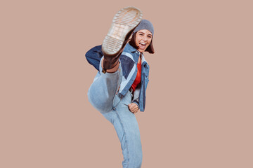 Cheerful beautiful woman is kicking the air, with leg up, looking at camera. Athletic girl making a direct kick wearing urban outfit. Photo of a young girl isolated on a beige background.
