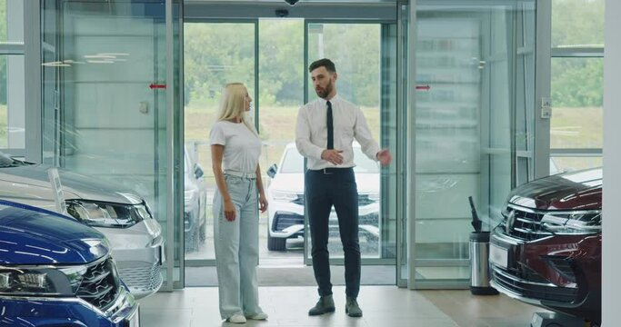 Female customer walking with salesman at modern salon and observing various luxury auto. Competent dealer helping young woman to make expensive purchase.