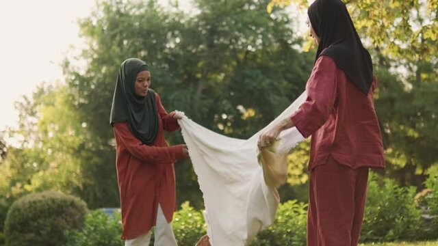 Two smiling Muslim women spreading out the picnic blanket outdoors