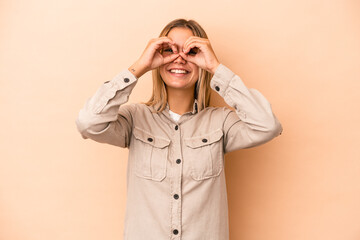 Young caucasian woman isolated on beige background showing okay sign over eyes