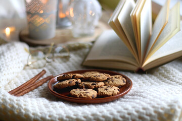 Plate of chocolate chip cookies, cinnamon sticks, soft blanket, open book and lit candles. Hygge at home. Selective focus.