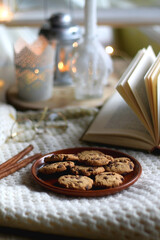 Plate of chocolate chip cookies, cinnamon sticks, soft blanket, open book and lit candles. Hygge at home. Selective focus.