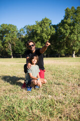 Obraz premium Loving father with disability talking with son. Man with mechanical leg in sunglasses and little boy sitting on grass in park, showing bird to son. Disability, family, love concept