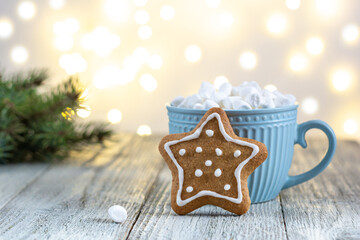 A blue cup of winter hot drink with marshmallows and gingerbread star on a white wooden background with bokeh.