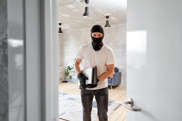Fototapeta na wymiar Thief with black balaclava stealing modern expensive laptop from apartment. Masked face. Man burglar stealing tv set from house