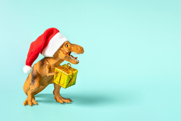 Dinosaur Rex in red Santa Claus hat holds golden gift box in its paws on blue background New Years...
