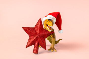 Dinosaur Rex in red Santa Claus hat holds star in its paws on pink background New Years Eve or...
