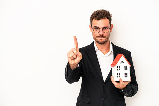 Young caucasian real estate holding a toy home isolated on white background showing number one with finger.