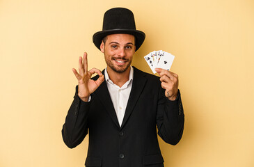 Young caucasian wizard holding magic card isolated on yellow background cheerful and confident showing ok gesture.