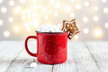 Obraz na płótnie Canvas A red cup of winter hot drink with marshmallows and gingerbread star on a white wooden background with bokeh.