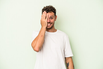 Young caucasian man isolated on green background having fun covering half of face with palm.