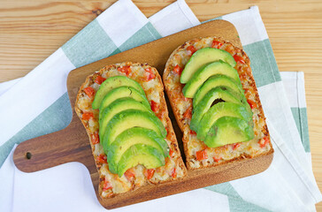 Pair of homemade grilled cheese toast topped with tomato and sliced avocado on wooden breadboard