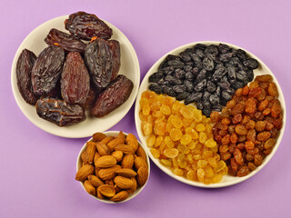 Dried fruits. Dried dates, light, brown, dark raisins on a round white plate, almonds in a cup on a lilac background. Top view. Close-up. Space for text.