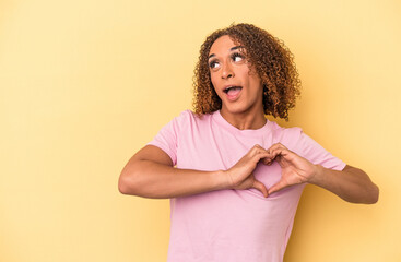 Young latin transsexual woman isolated on yellow background smiling and showing a heart shape with hands. - obrazy, fototapety, plakaty