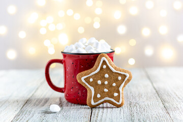 A red cup of winter hot drink with marshmallows and gingerbread star on a white wooden background with bokeh.
