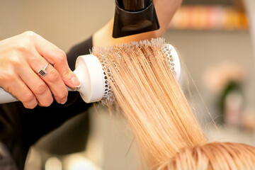 Naklejka premium Professional hairdresser dry hair with a hairdryer and round hairbrush in a beauty salon