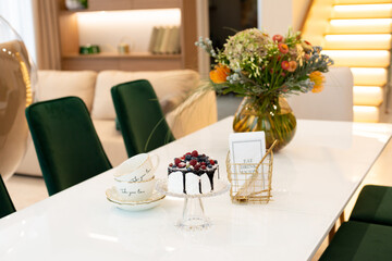 An exquisite berry cake on a glass stand on a table with a vase of flowers and a tea set in a modern designer interior. Soft selective focus, art noise