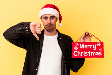 Young caucasian man holding a Merry Christmas placard isolated on yellow background showing a dislike gesture, thumbs down. Disagreement concept.