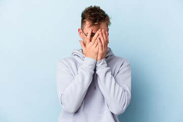 Young caucasian man isolated on blue background blink at the camera through fingers, embarrassed covering face.