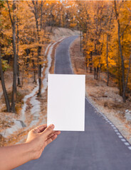 hand holding empty white sheet of paper. autumn background with road on a hill. sunny autumn day.
