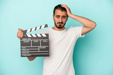 Young caucasian actor man holding clapperboard isolated on blue background being shocked, she has remembered important meeting.