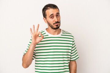 Young caucasian man isolated on white background winks an eye and holds an okay gesture with hand.